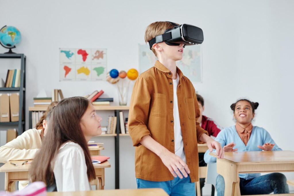 alt="Group on young students in a classroom, one wearing a virtual reality goggle where the others watched, a representation of education galaxy"