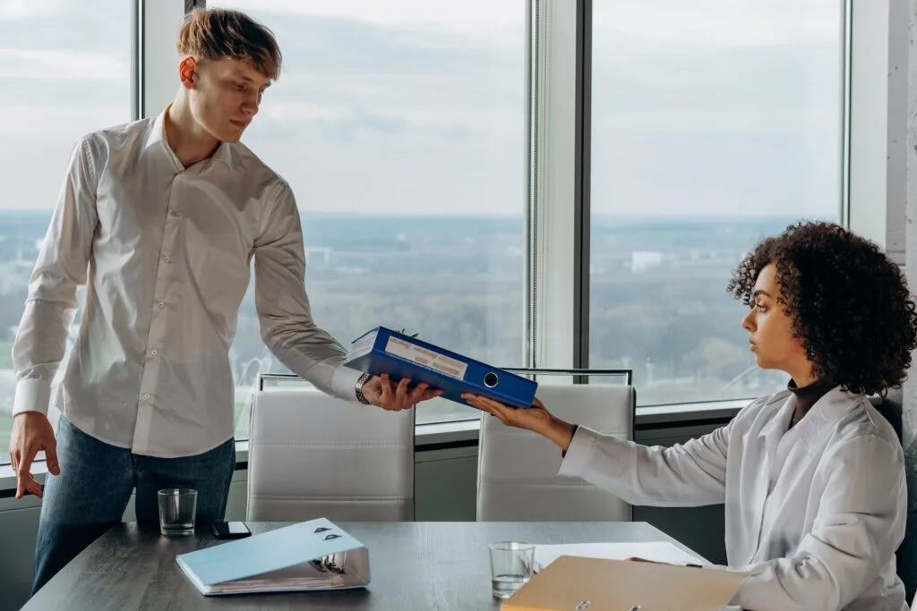 alt=" a young man standing and a young woman sitting behind a desk exchange affordable education books"