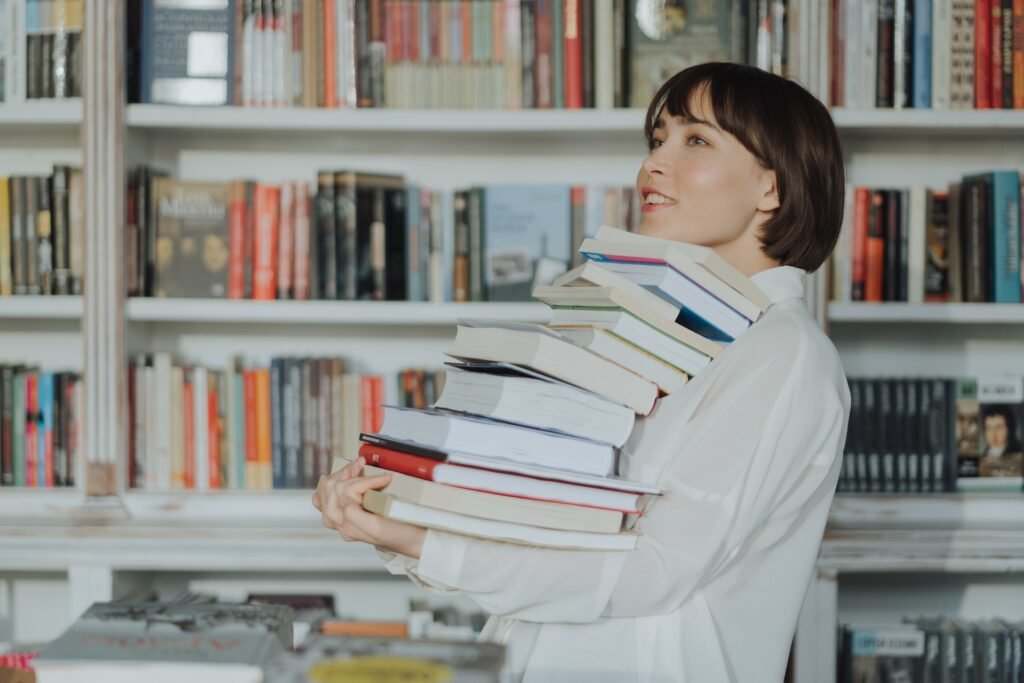 alt="a young woman carrying stacks of affordable education books in front of a book shelve filled with books"