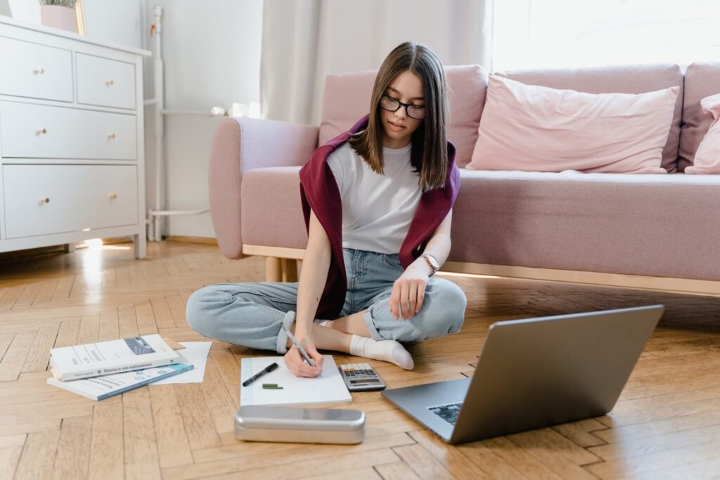 alt="a young student sitting on the floor at home in front of a pink couch with an open laptop and books having an alternative education session"