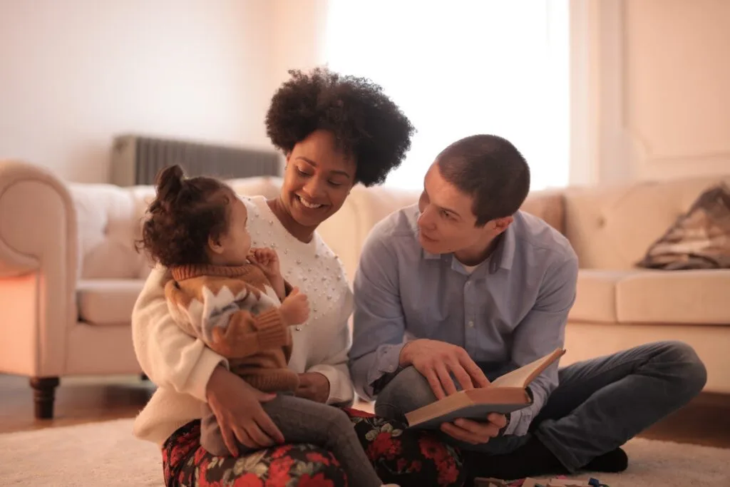 alt="a mother father and young daughter sitting on the floor with an open book having an alternative education session"