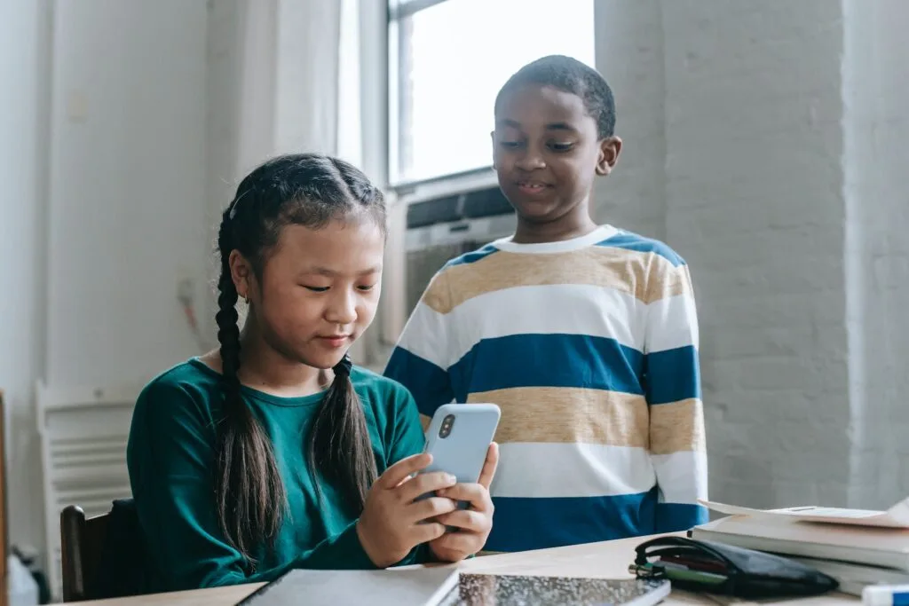 alt="a young male student and a young female student focusing on a cellphone with notebooks and on a table in front of them"