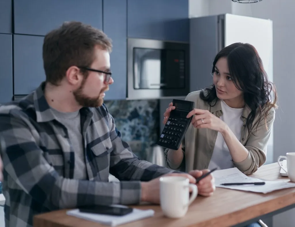 alt="a couple sitting next to each other looking at a calculator  with papers on the table in front of them"