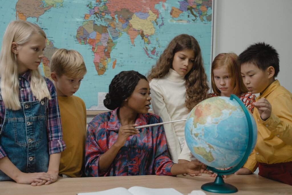 alt="a teacher sitting and pointing at a globe surrounded by her students standing around her"