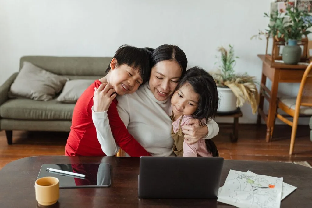 alt="a mother and her two children holding each other in front of an open laptop, a tablet and some papers on a table"