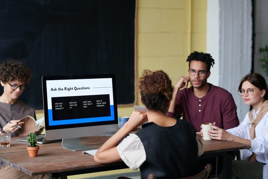 alt="a group of young people sitting around a table with a desktop pc displaying the words 'Ask the Right Questions' representing education galaxy".