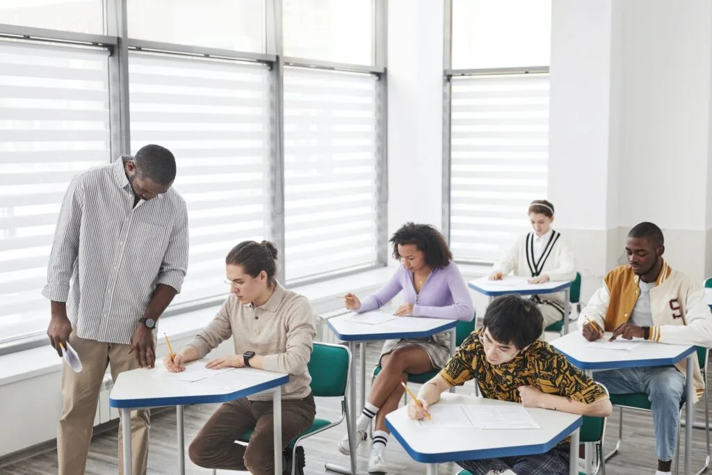 alt="Group of students taking an exam in a classroom monitored by a teacher as part of education dynamics"