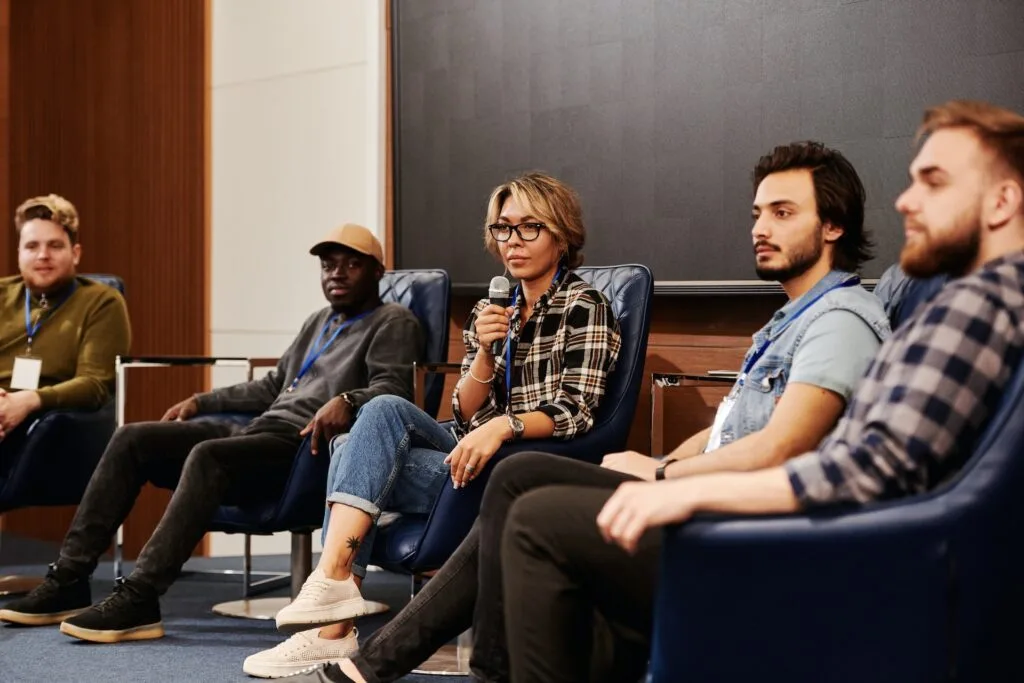 alt="A woman with a microphone with 3 males sitting on a stage part of education icon"