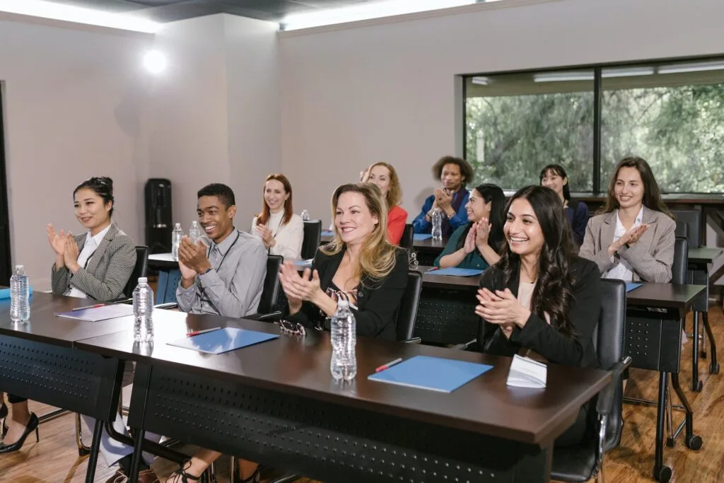 alt="A group of men and women sitting next to each other networking, smiling, and clapping part of education icon"