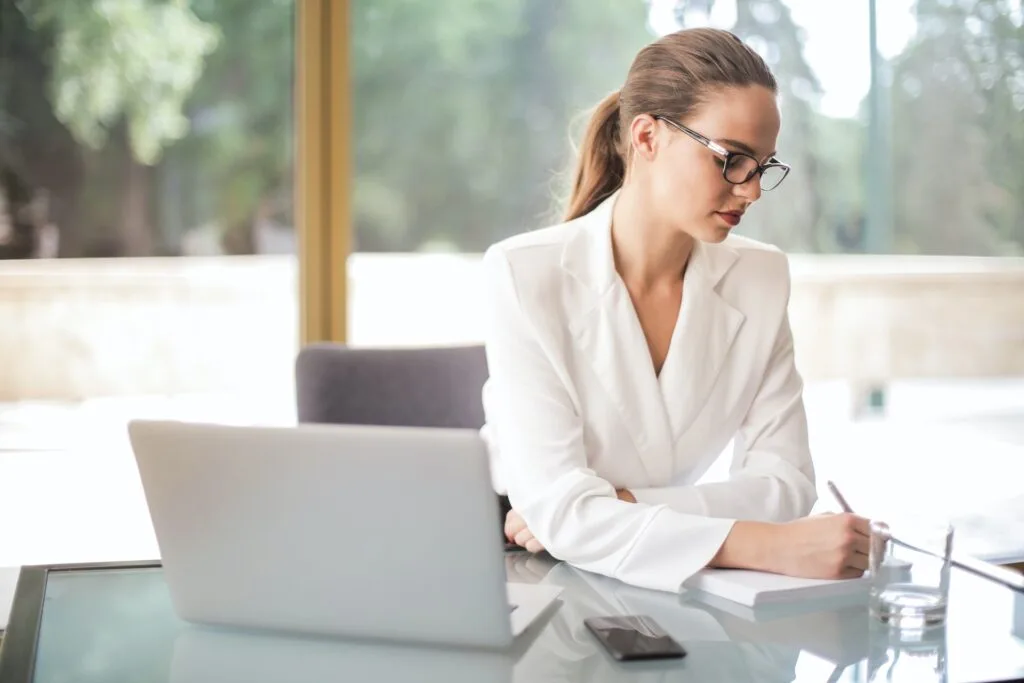 alt="A young woman writing on a piece of paper in front of a computer developing education background strategies"