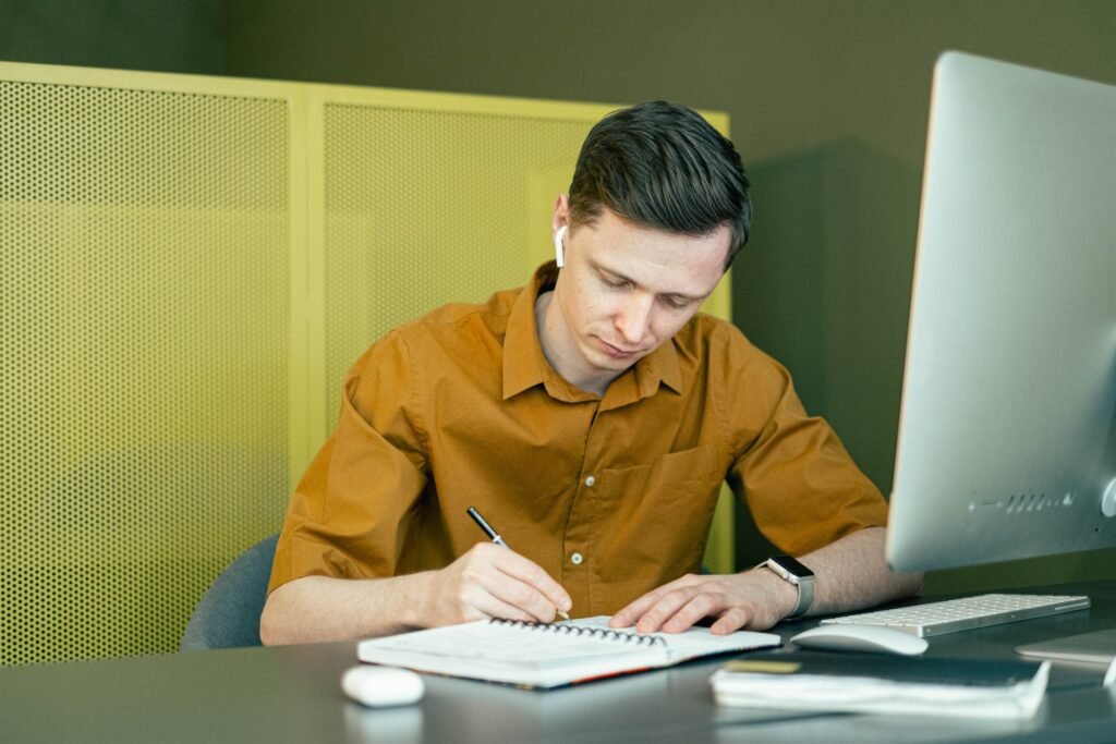 alt="a young man writing on a notebook in front of a computer exploring education background"