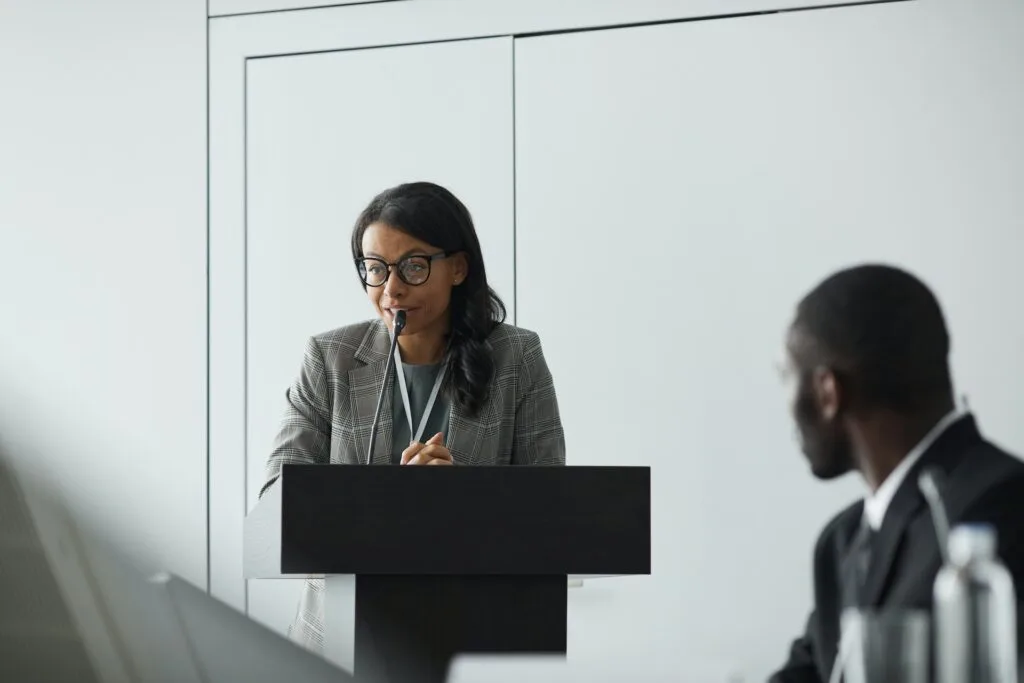 alt="A woman speaker at a podium speaking to a group as part of education icon"