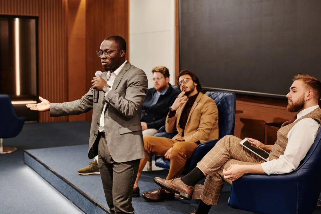 alt="A young male speaker standing with a microphone with a panel of other male speakers behind him part of education icon"
