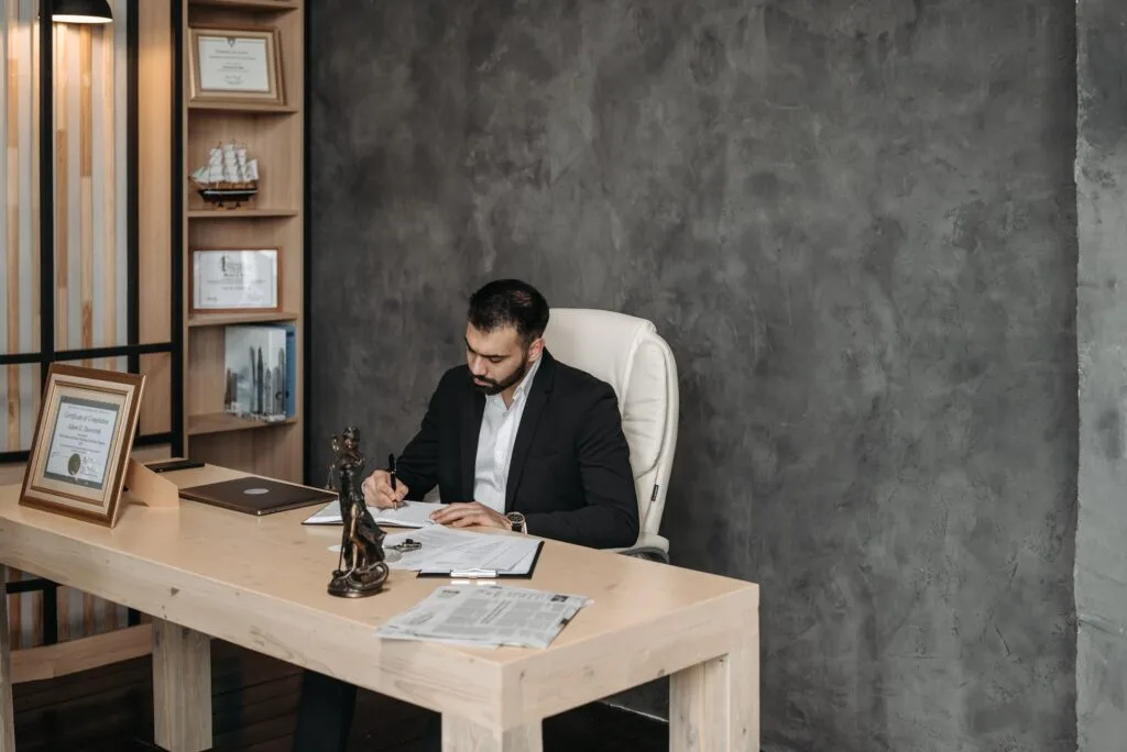 alt="A man sitting behind a desk writing on a notebook with a certification in a frame on the desk part of education background"